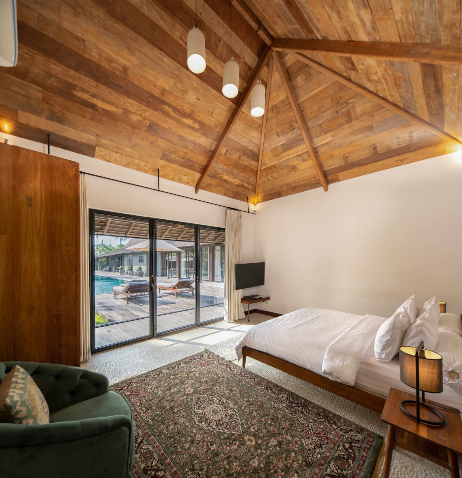 A pool house bedroom beneath its pitched teak ceiling, with doors onto the pool